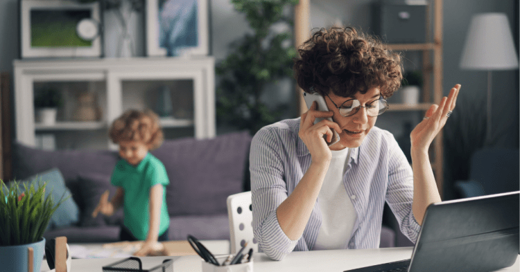 Woman sat at desk looking stressed. Talking on the phone. laptop in front of her and child in the background. Home setting.